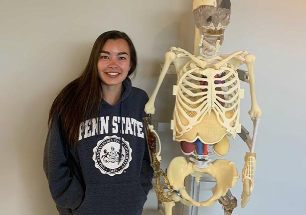 A graduate student wearing a Penn State hoodie stands next to a faux skeleton with a 3D-printed rib plate implant.