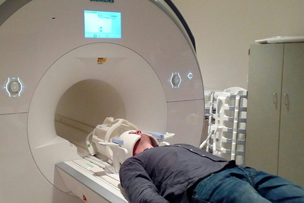 a person wearing jeans, gray button-down shirt, and ceramic helmet about to enter MRI chamber