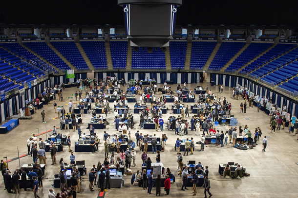 Photo taken from stands shows nearly eighty display tables set up in a large arena surrounded by people presenting and talking.