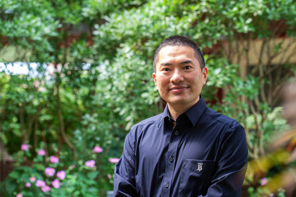 A researcher wearing a black button-down shirt smiles for a portrait in front of greenery
