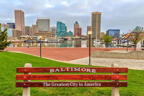 A city bench with the words, "Baltimore, the Greatest City in the Country" is seen before a city park, body of water, and city skyline.