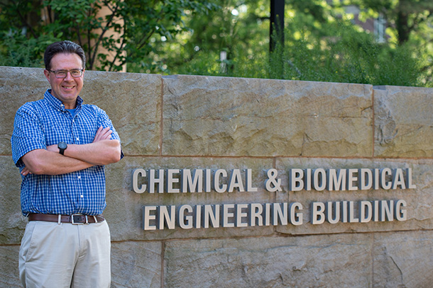 A person standing outside smiles at the camera. Behind him is a sign reading "Chemical Engineering and Biomedical Engineering Building."