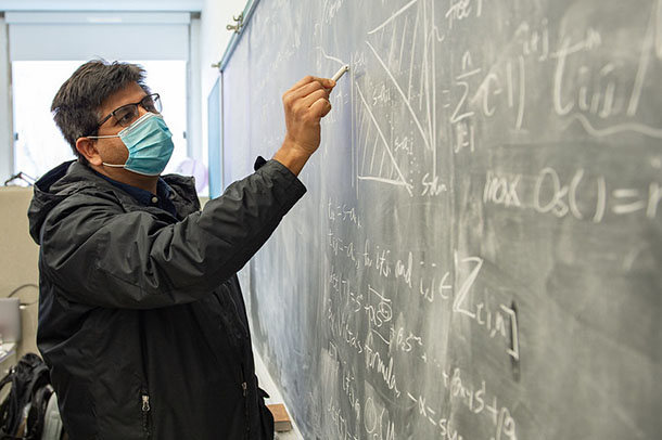 A man wearing a mask writes on a chalkboard.