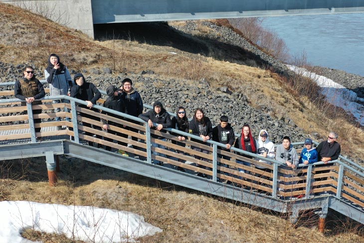 14 people pose on outdoor stairs near a river