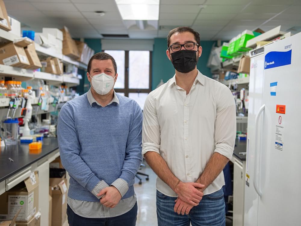 Two men wearing masks pose together in a lab, with lab equipment and a refrigerator in the background.