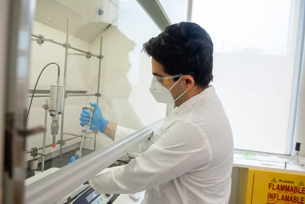 A person in a white lab coat, N95 mask and blue gloves uses a syringe-type device in a test tube.