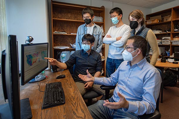 Five researchers wearing masks stand and sit around a computer, where two maps of the United States are displayed on the monitor. 