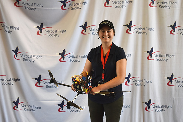 A person wearing a hat and a polo displays a drone in front of a backdrop that says "Vertical Flight Society."