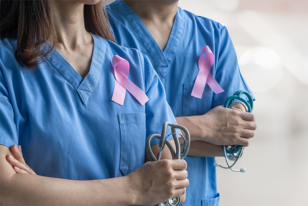 two individuals wearing medical scrubs and pink breast cancer ribbons pins hold stethoscopes with arms crossed