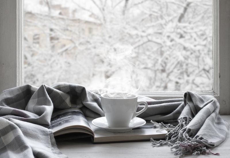 a steaming cup of coffee sits on a desk in front of window that looks out on a snow-covered landscape