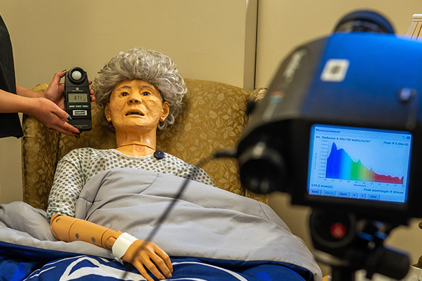 Dummy of elderly person wearing a monitor and surrounded by equipment is used for light experiment.