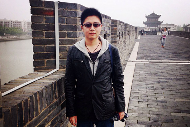 A person in sunglasses and a light jacket poses in front of a brick wall on a gray day. Buildings with traditional Chinese and with modern architecture can be seen in the background. 