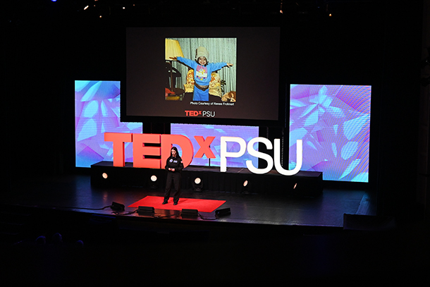 Renee Frohnert stands on stage with the TEDxPSU logo behind her and a screen hanging above her with a photo of her as a child. 