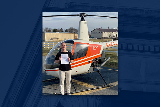 Young woman in light pants and dark shirt poses in front of orange and white helicopter on airfield.