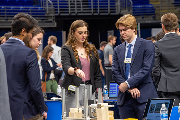 Young adults in business attire gather around an engineering exhibit.