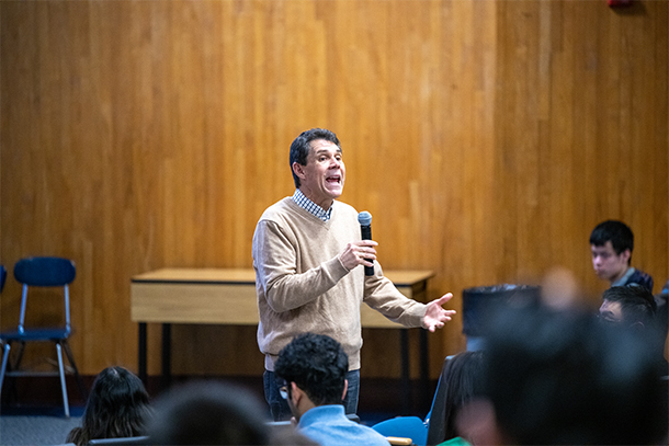 An individual holding a microphone speaks to a large crowd of students in an auditorium