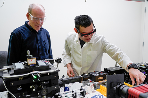 Two individuals wearing lab glasses adjust a microscope in a lab. 