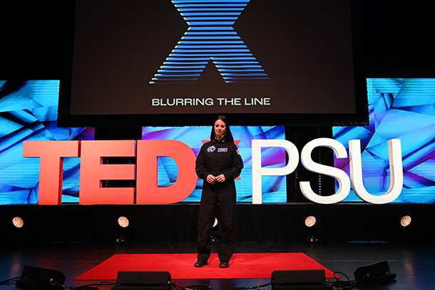 Renee Frohnert stands on stage with the TEDxPSU logo behind her and a screen hanging above her with "Blurring the Line" on it. 
