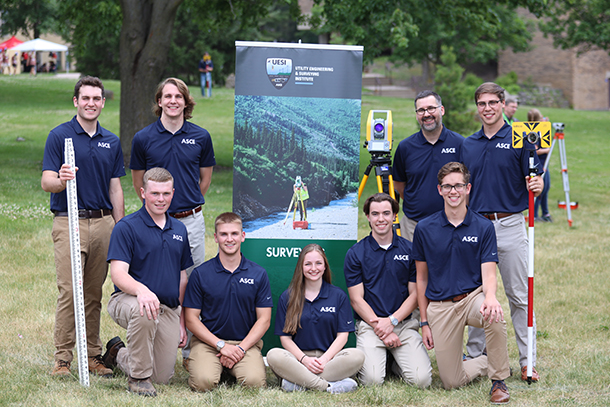 A group of students wearing navy blue polo shirts poses outdoors with surveying equipment.