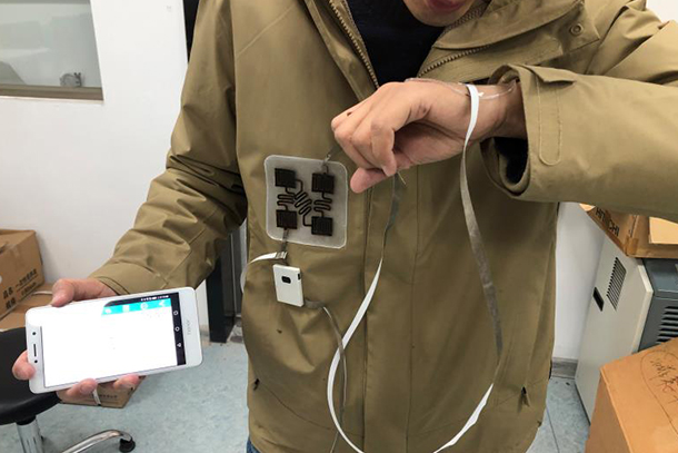 A person in a tan jacket breathes onto their wrist. A wire connects the wrist sensor to a monitor on the jacket. The person holds a phone in their hand, showing data is collected.