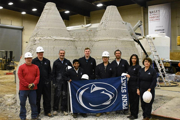 Nine people in hard hats holding a Penn State flag stand in front of 3D printed structures
