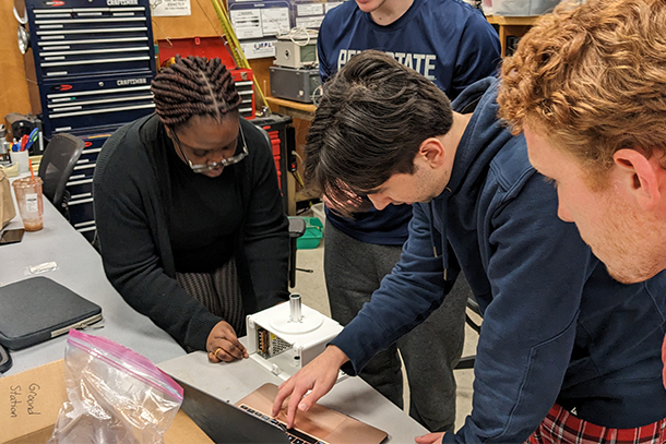 Four students work at a laptop and on a small, white, rectangular piece of equipment