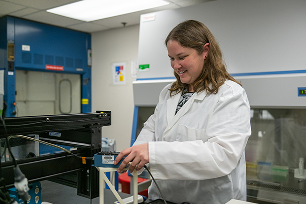 A person in a white lab coat smiles and adjusts a blue rectangular piece of equipment