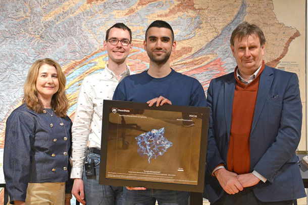 four people smile for photo in front of map. Person in center of photo holds a printed image of the header image.