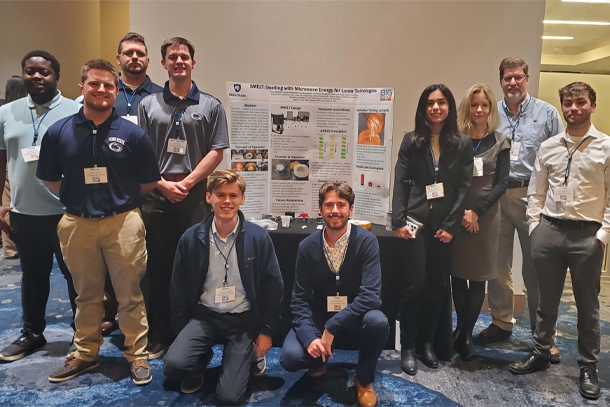 A team of 10 people, dressed in business casual clothing, pose for a photo in front of a project poster.