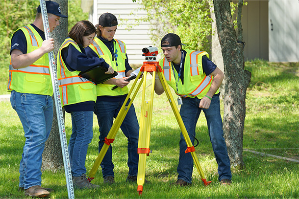 Four people in neon vests consult clipboards and use a piece of surveying equipment.