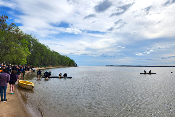 People stand on the shore of a lake while canoes launch from the shore.