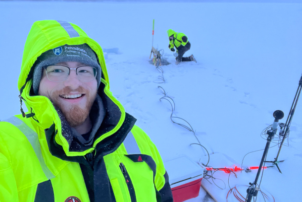 An individual wearing a neon yellow jacket and thermal hat stands on a snow-covered lake. In the background, a researcher takes measurements. 