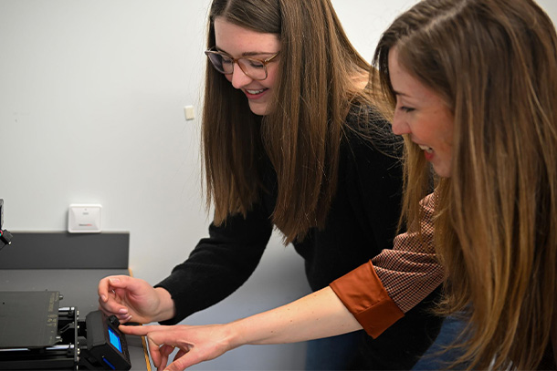 two people smile while pointing towards a small touchscreen of a 3D printer.