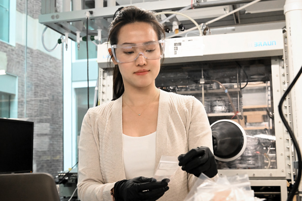 An individual in a lab wearing protective glasses and gloves handles a sample of a coin cell battery.