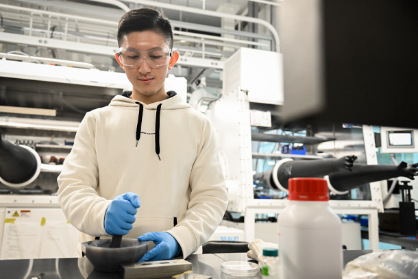 An individual in a lab wearing protective glasses and gloves uses a mixing bowl to combine a sample. 