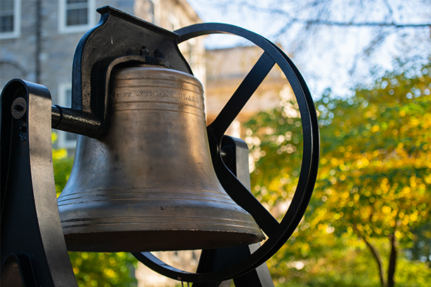 Old main's bronze bell on display in front of green foliage.