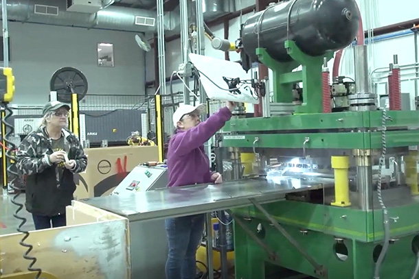 Two people in baseball caps and safety glasses work in a manufacturing plant.