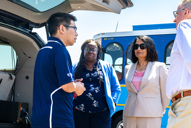  Four people talk in a group in front of a blue bus and next to a vehicle with an open truck. 