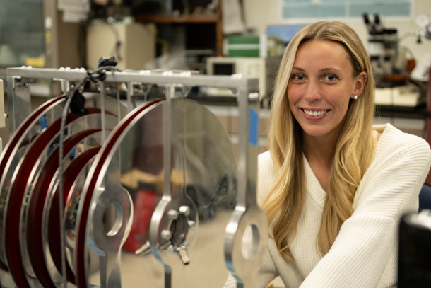 A portrait of an individual wearing a white sweater using lab equipment in a lab setting. 