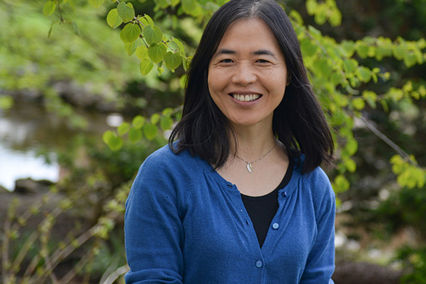 An individual wearing a blue sweater in front of greenery. 