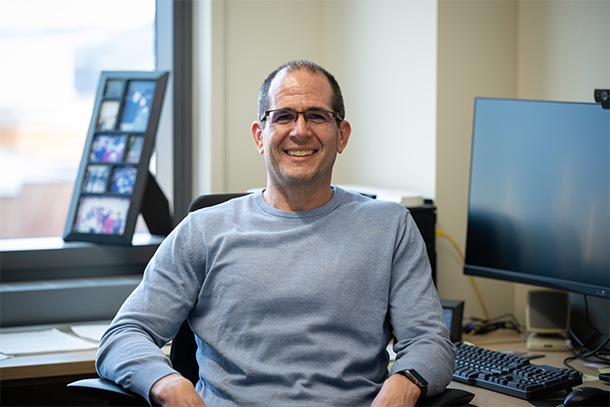 A person in glasses sits at a desk with a computer monitor in the background 