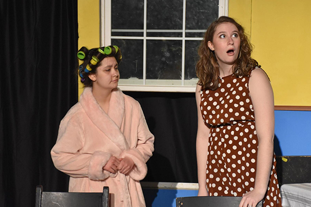 An individual in a bathrobe and one in a polka dotted dress stand in front of a window