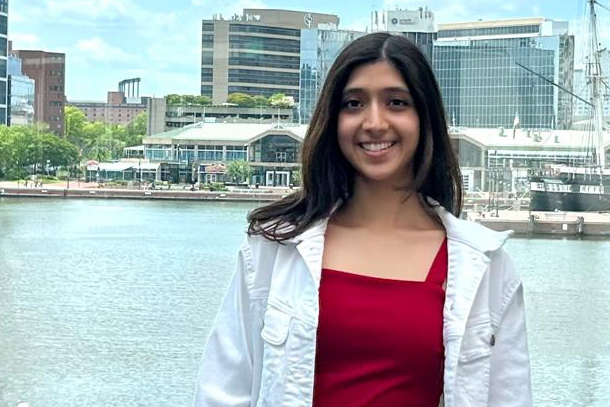 A person in a red dress and white jacket stands in front of a body of water with tall buildings in the background.