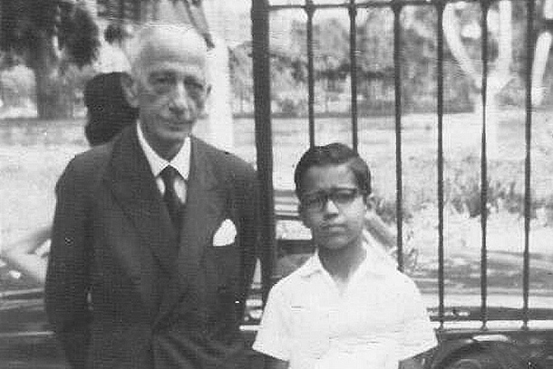 A black and white image of an adult and child posing for the camera in front of a gate and a car. 