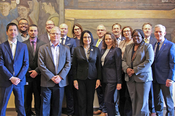 A group of business professionals stand and smile for group photo in front of mural.