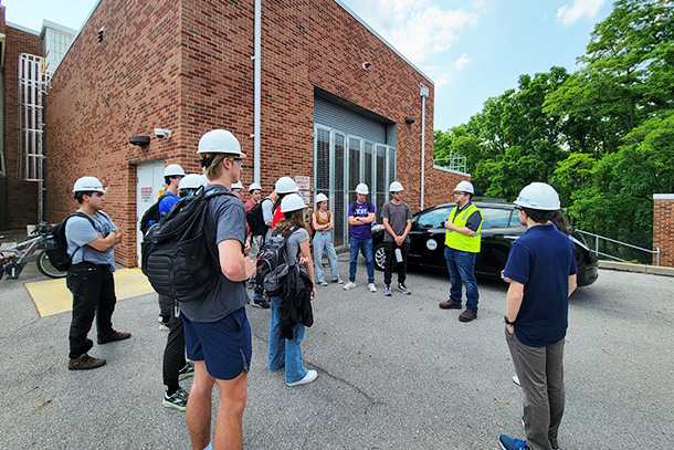 Students in hard hats stand outside of a steam plant building listening to an instructor in a neon vest.  