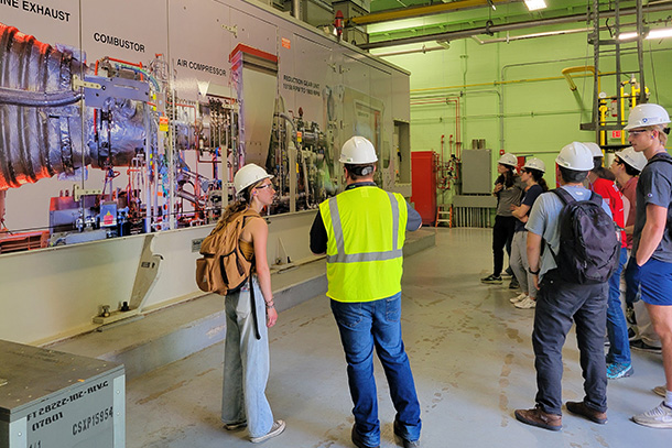 Students in hard hat and instructor in hard hat and neon vest look at a large rendering of a gas turbine engine shown on a wall in a steam plant building. 