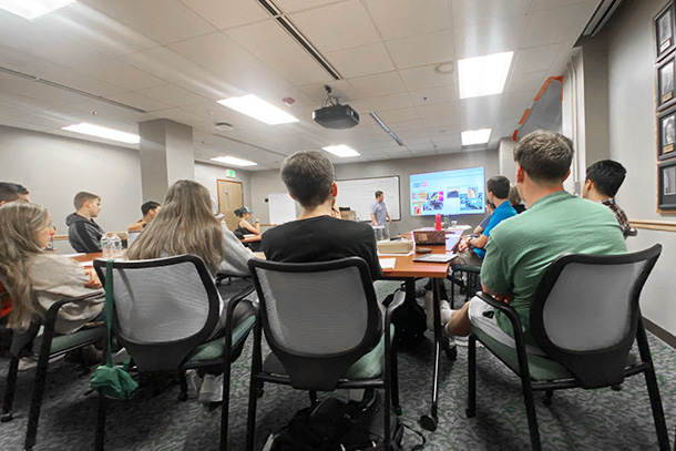 Students sit in a classroom and listen to an instructor at the front of the class presenting on a projector screen. 