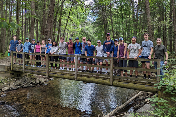 Students and faculty members stand on a small bridge, surrounded by woods during a hike. 