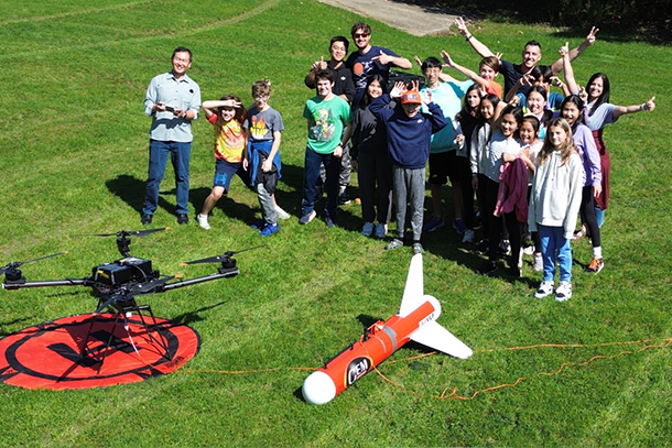 Group of children and adults standing behind a large drone in a field.